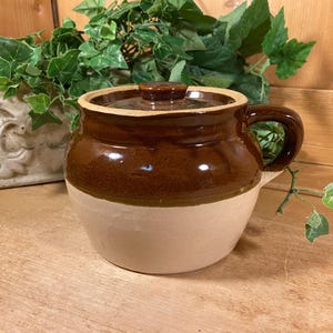 May include: A two-toned ceramic crock with a glossy dark brown glazed upper section and a matte beige lower section. The pot features a single side handle, a matching brown lid with a small central knob, and a rounded, wide-bodied shape.