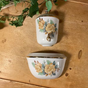 May include: A white ceramic water dispenser and soap dish set. The dispenser has a gold-colored spigot and lid. Both pieces are decorated with yellow roses and gold trim. The set is on a wooden surface.