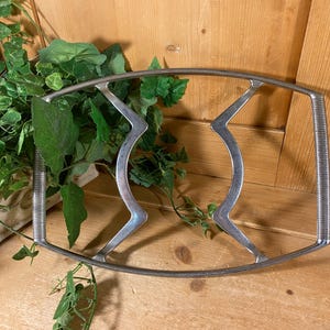 May include: A silver-toned metal trivet with a rectangular shape and curved, zigzag design in the center. The trivet has ribbed handles on each end. Green leafy plants are in the background.