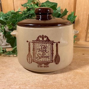 May include: Tan ceramic kitchen canister with a brown lid and handles. The canister features a brown graphic with the words "The Fork & Spoon" and the date "1776-1976". The canister is sitting on a wooden surface.