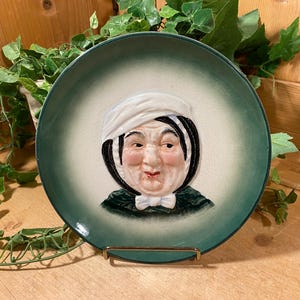 May include: A decorative ceramic plate with a portrait of a woman wearing a white bonnet and a bow tie. The plate has a green border and a cream-colored center. The plate is displayed on a gold-colored stand.