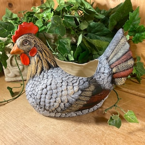 May include: A ceramic chicken-shaped planter featuring a textured grey and white body with brown and red accents on the wings and tail. The head has a bright red comb and wattle with a yellow beak. The open top allows for planting or storage.