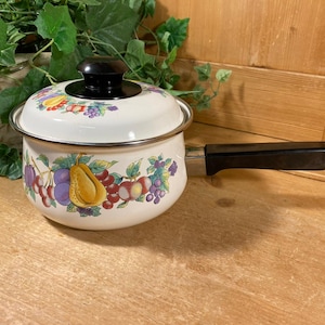 May include: Cream-colored enamel saucepan with a black handle and lid. The pot is decorated with a colorful fruit pattern, including grapes, pears, and plums. A vintage kitchenware item.