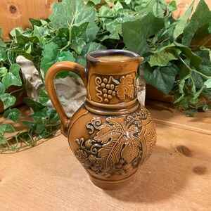 May include: A brown ceramic pitcher with a vine and grape design. The pitcher has a handle and a spout. The pitcher is decorated with a brown glaze.