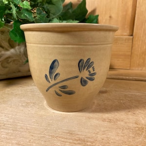 May include: A tan stoneware crock featuring a tapered shape and a wide rim. The exterior is decorated with a hand-painted blue floral motif consisting of a single stem with leaves and a stylized flower blossom centered on the front of the vessel.