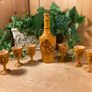 May include: A decorative wooden liquor set featuring a tall bottle with a stopper and six small shot glasses. The set is painted in a warm yellow color with brown floral designs. The shot glasses and bottle are approximately 3 inches tall.