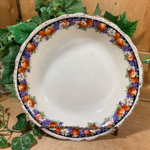 May include: A white ceramic serving bowl with a scalloped edge, decorated with a border of orange fruit, purple flowers, and white blossoms. The bowl is displayed on a decorative metal stand.