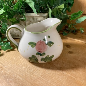 May include: A white ceramic creamer with a green rim and floral design. The creamer features a pink flower with green leaves and a white background.