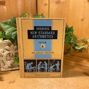 May include: A vintage Grade 6 arithmetic textbook titled "Iroquois New Standard Arithmetics." The book has a tan cover with blue and black text and illustrations. The cover reads "Enlarged Edition" and "Patton and Young."