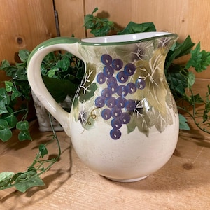 May include: Cream-colored ceramic pitcher with a large handle and a spout. The pitcher is decorated with a cluster of purple grapes and green leaves. The rim and handle are trimmed in green. The pitcher is sitting on a wooden surface.