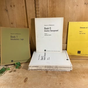 May include: A collection of vintage math textbooks, including "Elements of Mathematics" books 0, 1, and 2. The books have white and colored covers with text. The books are stacked on a wooden surface.