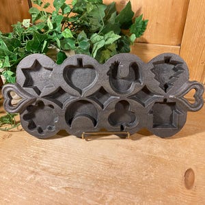 May include: A dark gray cast iron cake pan featuring eight distinct molded cavities in various shapes including a star, heart, rooster, tree, flower, crescent moon, clover, and house, with heart-shaped handles on both ends of the rectangular frame.