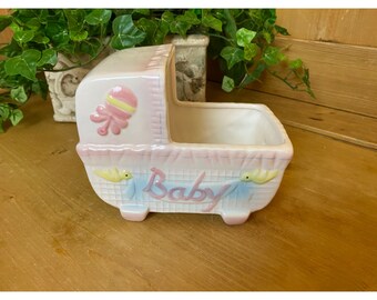 May include: A white ceramic baby crib planter with pink and blue accents. The crib has a yellow bird on each side and the word "Baby" written across the front.