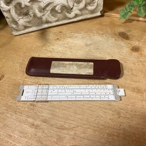 May include: A white plastic slide rule with a clear sliding cursor and metal end caps, accompanied by a dark brown leather protective case featuring a rectangular label with the text "FABER-CASTELL" and additional printed information.