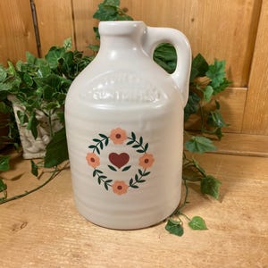 May include: A white ceramic jug with a handle and a narrow neck. The front features a printed design of a dark red heart surrounded by a circular wreath of green leaves and four light orange flowers. The jug has a smooth, glossy finish.