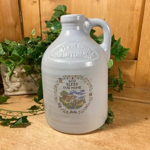 May include: A light blue ceramic jug with a handle, featuring the text "Clinton China" and a circular emblem that reads "God Bless Our Home." The jug is surrounded by greenery and sits on a wooden surface.