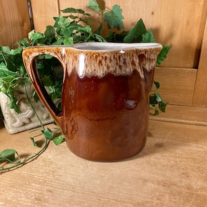 May include: A ceramic pitcher featuring a glossy, deep brown glaze on the lower body that transitions into a creamy, off-white drip pattern around the rim. The vessel includes a sturdy, curved handle and a wide, functional pouring spout.