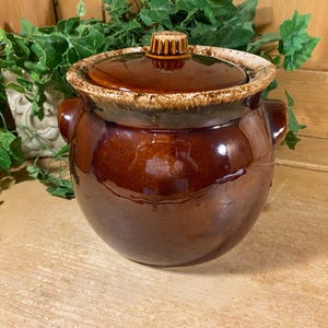 May include: A vintage brown ceramic bean pot with a lid. The pot has a glossy finish and a two-tone design, with a lighter rim and a darker body. It features two side handles and a knob on the lid.
