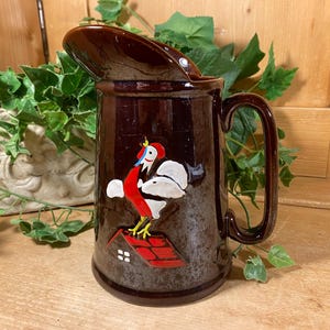 May include: A dark brown ceramic pitcher with a hinged lid and a large handle. The pitcher features a colorful rooster design on the front, with red, white, and yellow accents. The pitcher is likely vintage and decorative.