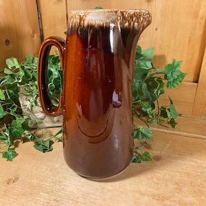 May include: A tall, glossy brown ceramic pitcher with a large handle and a pouring spout. The top edge has a textured, light brown and white glaze. The pitcher is set against a wooden background with green foliage.