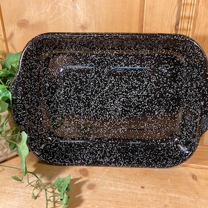 May include: A rectangular black enamel roasting pan with white speckles. The pan has rounded corners and handles on each side. The pan is empty and sits on a wooden surface.