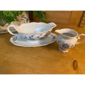 May include: A white gravy boat with a blue floral pattern and a matching saucer. The gravy boat has a handle and a spout. The saucer is oval shaped. There is also a small white creamer with a blue floral pattern.