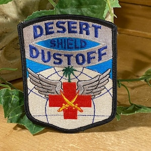 May include: Embroidered patch with the words "DESERT SHIELD DUSTOFF" in blue lettering. The patch features a red cross with crossed swords, wings, and a palm tree. The background is a light blue and white design.