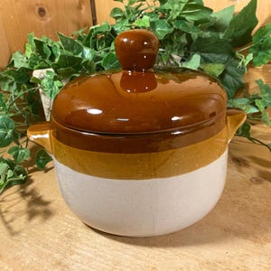 May include: A round ceramic casserole dish with a lid, featuring a tri-color design with a dark brown top, a mustard yellow middle band, and a cream-colored base. The pot includes two side handles and a knob on the lid for easy lifting.