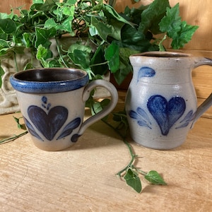 May include: A ceramic mug and a matching pitcher, both featuring a light gray speckled glaze with hand-painted dark blue heart motifs and leaf accents. The mug has a dark brown interior rim, while the pitcher has a curved handle and spout.