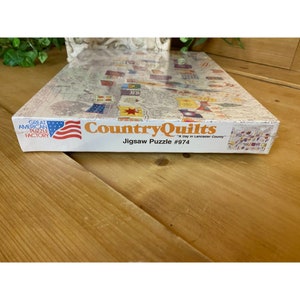 Może przedstawiać: Pudełko do układanki przedstawiające kolorową ilustrację patchworkowych kołder z tekstem "Country Quilts" i "A Day in Lancaster County". Pudełko zawiera r&oacute;wnież tekst "Great American Puzzle Factory" i "Jigsaw Puzzle #974".
