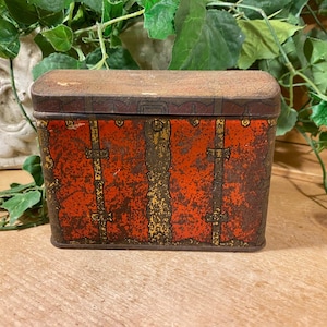 May include: An antique rectangular tin box with a rusty, distressed finish. The box features a red background with gold-colored vertical accents and decorative clasps. The lid is a darker brown color.