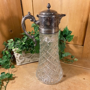 May include: A vintage glass pitcher with a silver-colored metal top, handle, and spout. The glass body features a diamond-cut pattern. The pitcher is displayed on a wooden surface with greenery in the background. A decorative piece for serving beverages.