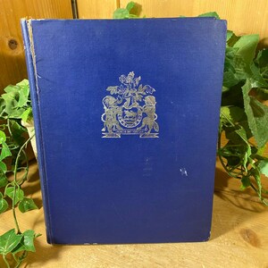 May include: A vintage book with a dark blue cover. The cover features a silver emblem with a crest and text. The book is slightly worn, with visible wear on the edges. Green vines are in the background.