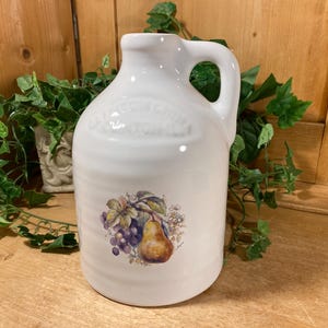 May include: White ceramic jug with a handle, featuring a fruit illustration of grapes and a pear. The jug has embossed text near the top. A decorative item for the kitchen or dining room.