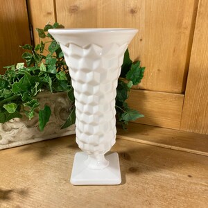 May include: A white milk glass vase featuring a repeating geometric honeycomb pattern across its exterior. The vase has a wide, flared circular rim that tapers down to a narrow neck, supported by a square pedestal base with a smooth finish.