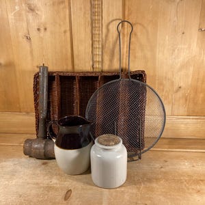 May include: A collection of kitchen items including a brown woven cutlery tray, a dark brown and cream ceramic pitcher, a white ceramic jar with a cork lid, a metal mesh splatter screen with a long handle, and a weathered wooden mallet.