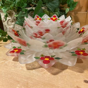 May include: A frosted glass decorative bowl in the shape of a flower. The bowl has multiple layers of petals, each adorned with red and yellow floral designs. The bowl is sitting on a wooden surface.