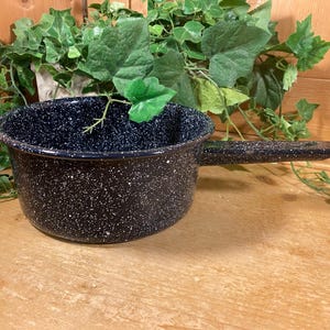 May include: A dark blue enamel saucepan with a speckled white pattern. The pan has a single handle and a rounded shape. The background includes green foliage and a wooden surface, suggesting a rustic kitchenware theme.