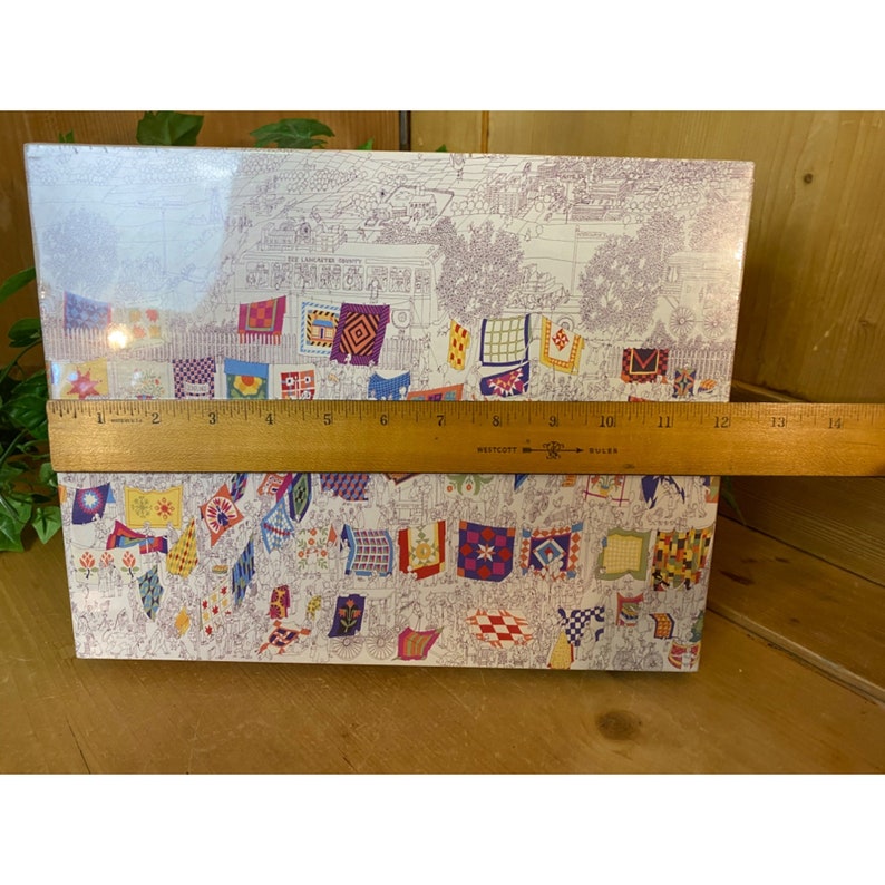 Może przedstawiać: Ilustracja w stylu vintage przedstawiająca plac miejski z kolorowymi patchworkowymi kocami wiszącymi na sznurkach do prania. Ilustracja jest wydrukowana na białym tle i oprawiona w drewnianą ramkę.