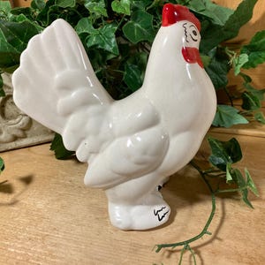 May include: A white ceramic chicken figurine with a glossy finish, featuring a bright red comb and wattle. The bird is depicted in a standing profile position with its tail feathers fanned upward. A black signature is visible on the base.
