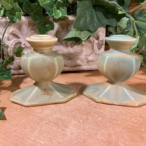 May include: Two vintage candle holders with a light green and beige color scheme. The candle holders have a bulbous body and a hexagonal base. The background includes greenery and a decorative planter.