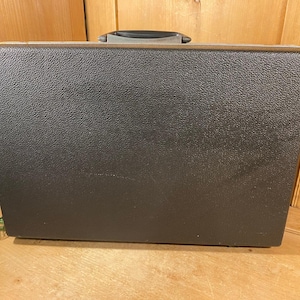 May include: A dark gray, textured briefcase with a black handle. The rectangular case has a slightly worn appearance, suggesting it has been used. The handle is a darker shade of gray. The briefcase is sitting on a wooden surface.