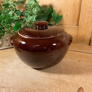 May include: A round, dark brown glazed ceramic pot with a matching lid and two side handles. The pot features a smooth, glossy finish and a circular knob on the center of the lid. The design is simple and functional with a deep, rounded body.