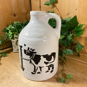 May include: A white ceramic jug with a handle, featuring a black graphic print of a cow tethered to a post. The cow is depicted in profile with a bell around its neck, and a small bird sits atop the post. The jug has a smooth, glossy finish.
