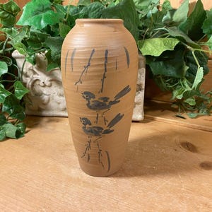 May include: A brown ceramic vase with a stylized design of two birds perched on branches. The vase has a smooth, slightly textured surface.