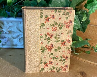 May include: A vintage book with a floral design on the cover. The cover is a light beige color with a darker beige border. The flowers are red, green, and white.