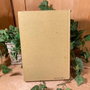 May include: A hardcover book with a light tan cover. The book is surrounded by green leafy vines and a decorative white pot. The spine of the book is visible, with text at the bottom.