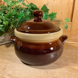 May include: A round ceramic bean pot with a glossy dark brown glaze on the bottom and lid, featuring a contrasting light beige band around the center. The pot includes two small side handles and a matching lid with a central knob handle.