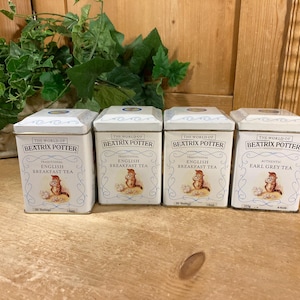 May include: Four white rectangular metal tea tins featuring The World of Beatrix Potter branding. Each tin displays a small illustration of a squirrel, blue decorative line work, and text identifying the contents as English Breakfast or Earl Grey tea.