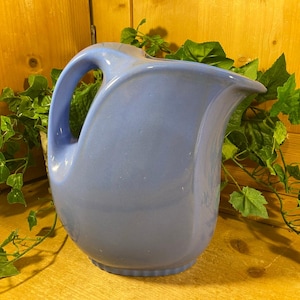 May include: A light blue ceramic pitcher with a curved handle and spout. The pitcher is set against a wooden background with green leafy vines. The pitcher has a smooth, glossy finish and a rounded base.
